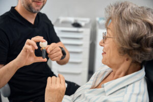 A senior woman in a dental office listens as a dentist shows her a dental implant model, illustrating how tooth replacement with implants works.